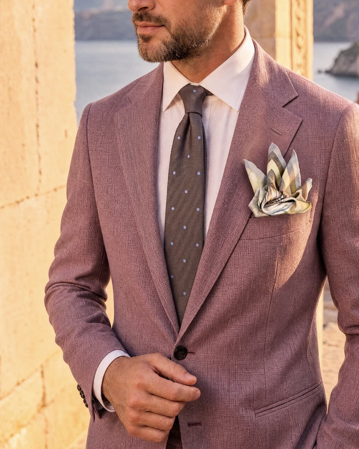 Cravatta in seta grenadine jacquard grigia a pois celesti indossata con camicia bianca e giacca in lino color malva, abbinata alla pochette da taschino Fluttuando nelle cromie minerali neutre, davanti al mare