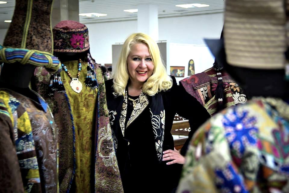 Aida Abdullaeva smiling among embroidered Uzbek robes, wearing Davide Cristofaro’s silk scarf