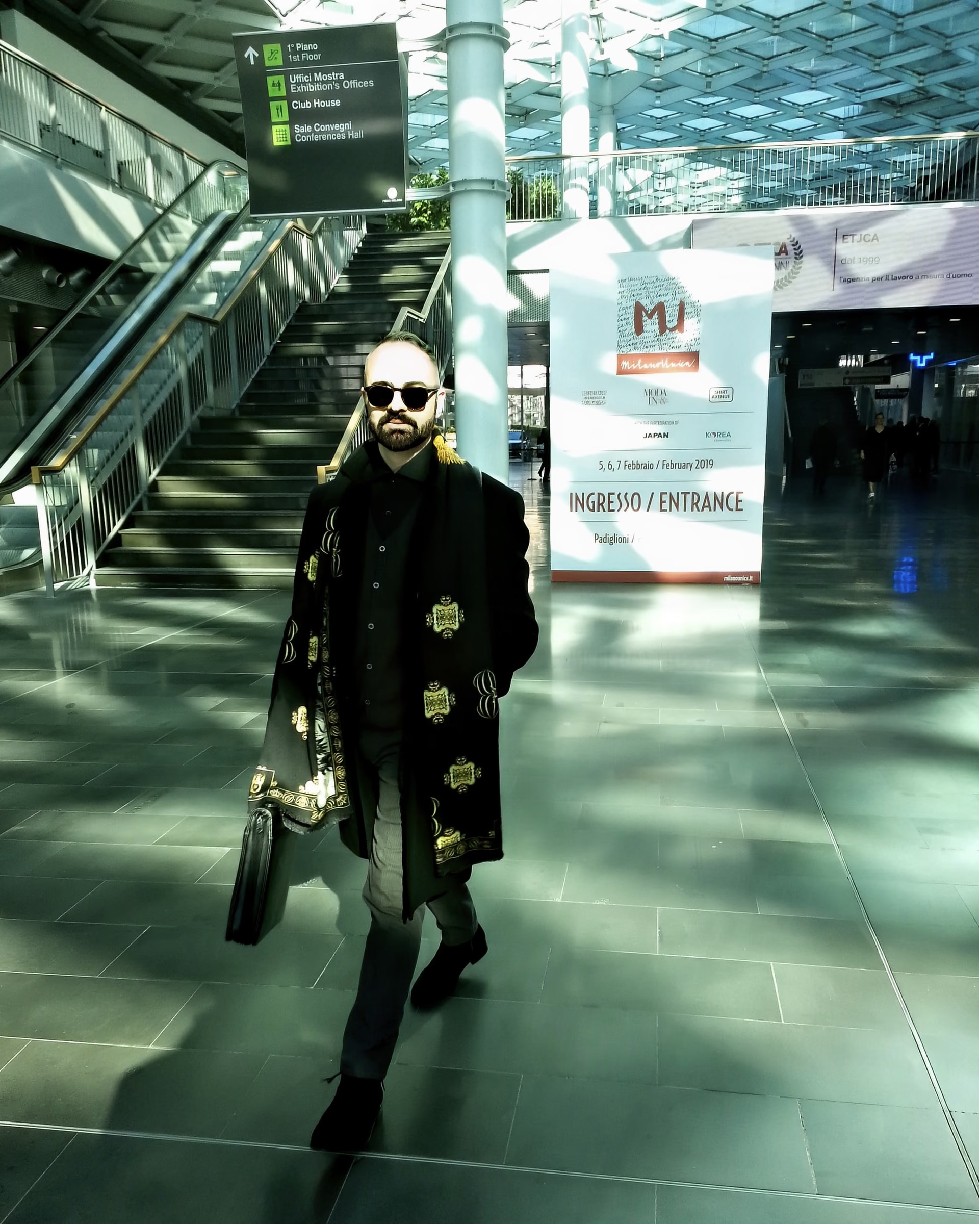 Davide Cristofaro en manteau noir à motifs dorés, sac à la main, traverse le hall d’entrée de Milano Unica 2019