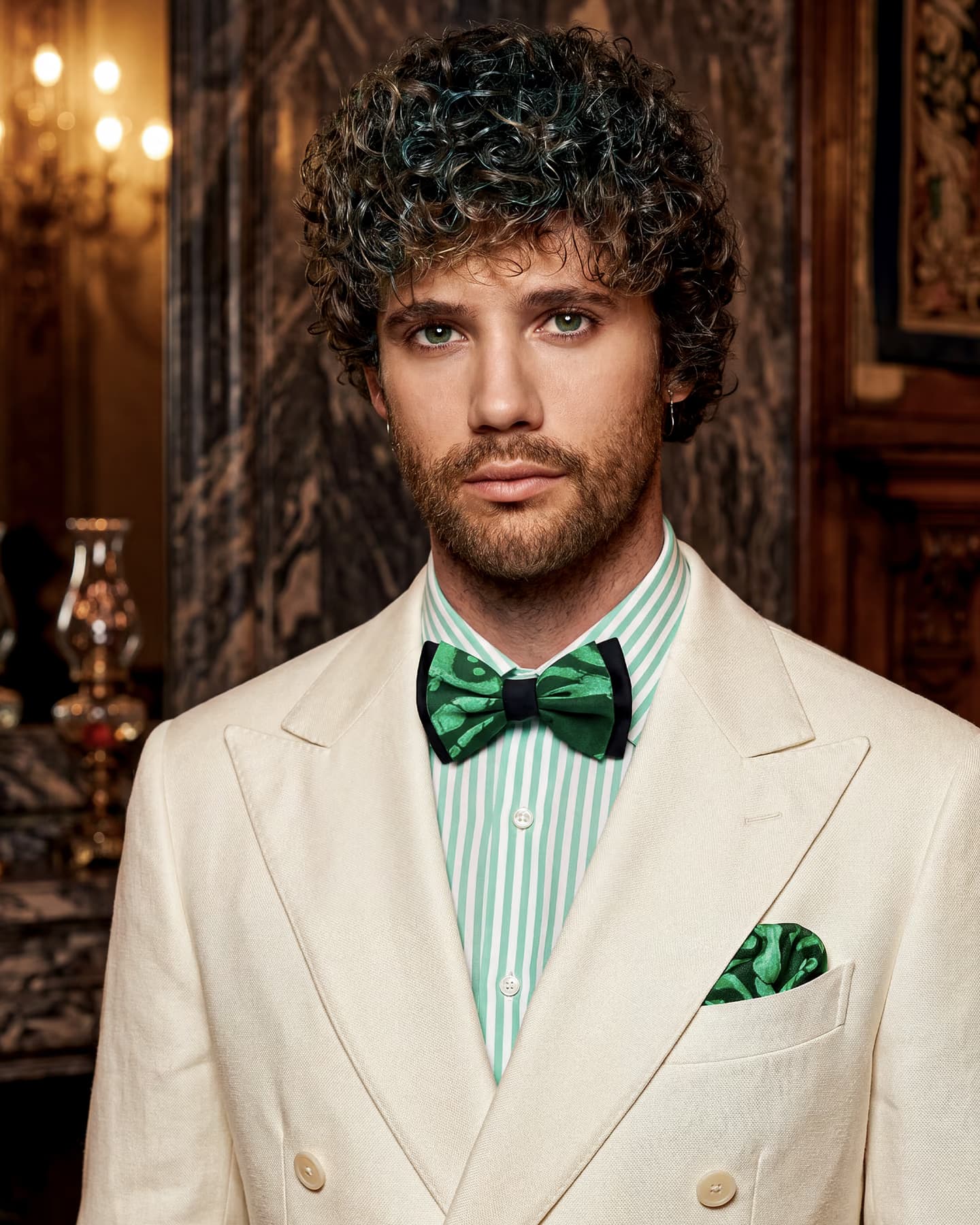 Modello con papillon in seta verde e nero stampa Liberty e pochette coordinata, giacca doppiopetto avorio e camicia a righe verdi, in un salone d'epoca