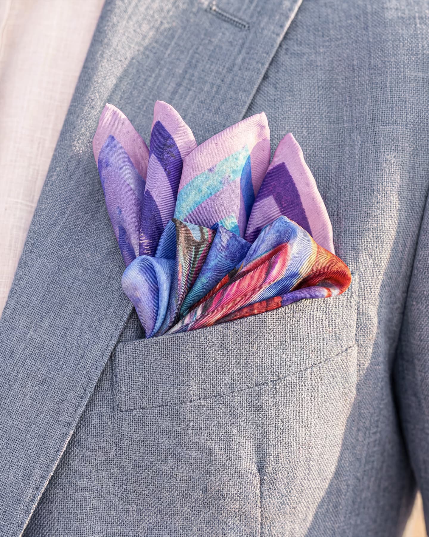 Dettaglio della pochette in twill di seta Fluttuando dalla stampa pittorica viola, lilla, azzurro e corallo, piegata a sbuffo nel taschino di una giacca in lino azzurro