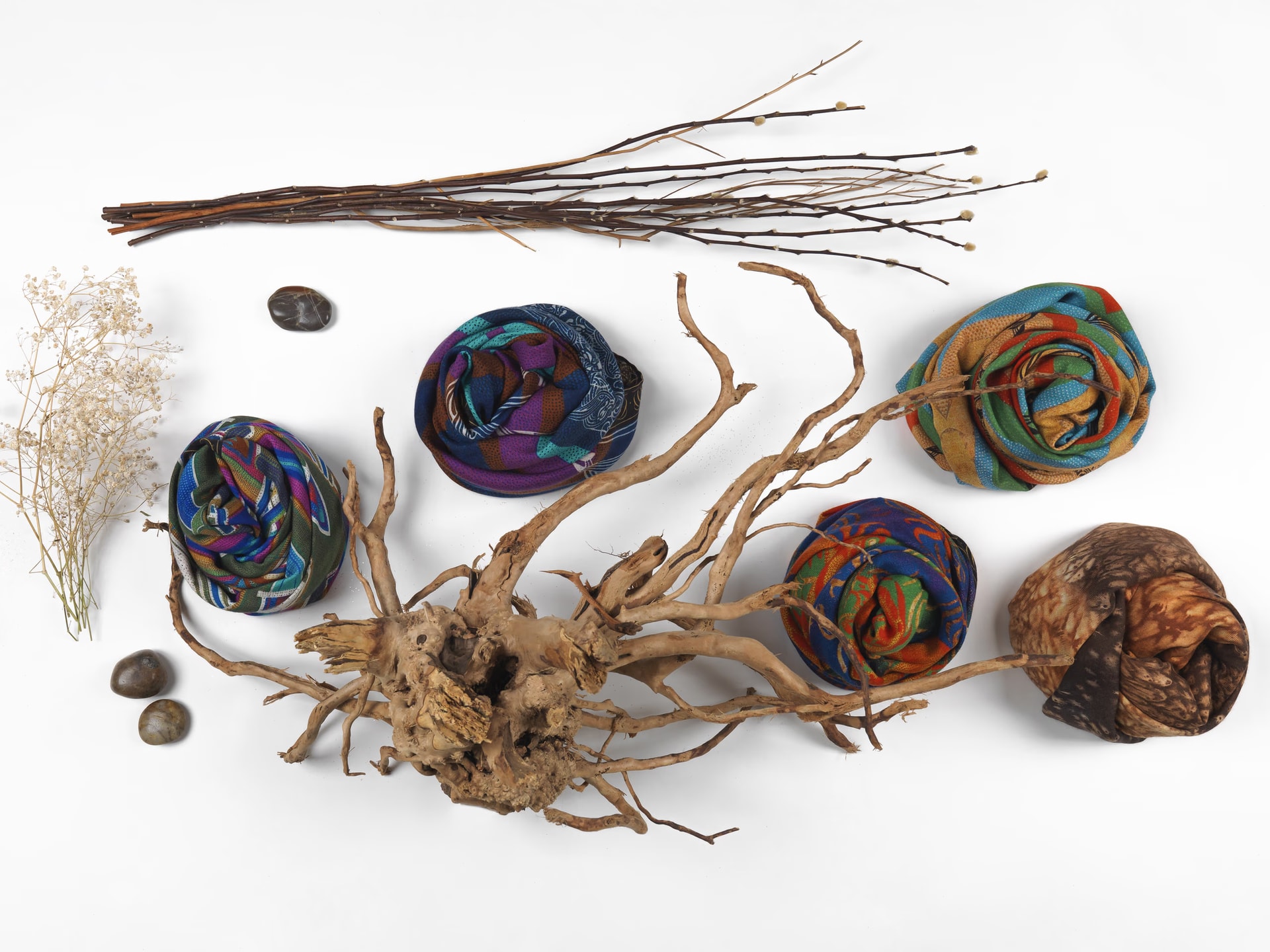 Cinque foulard arrotolati dai colori intensi disposti intorno a una radice di legno, con rami secchi, sassi e fiori secchi