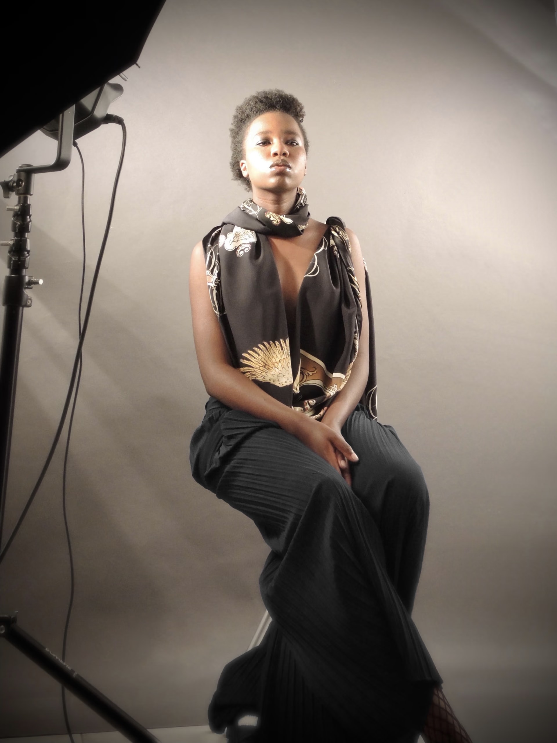 Backstage: Esther Ngumo seduta su uno sgabello in studio con un foulard di seta nero e oro e pantaloni plissettati, accanto a una luce da set