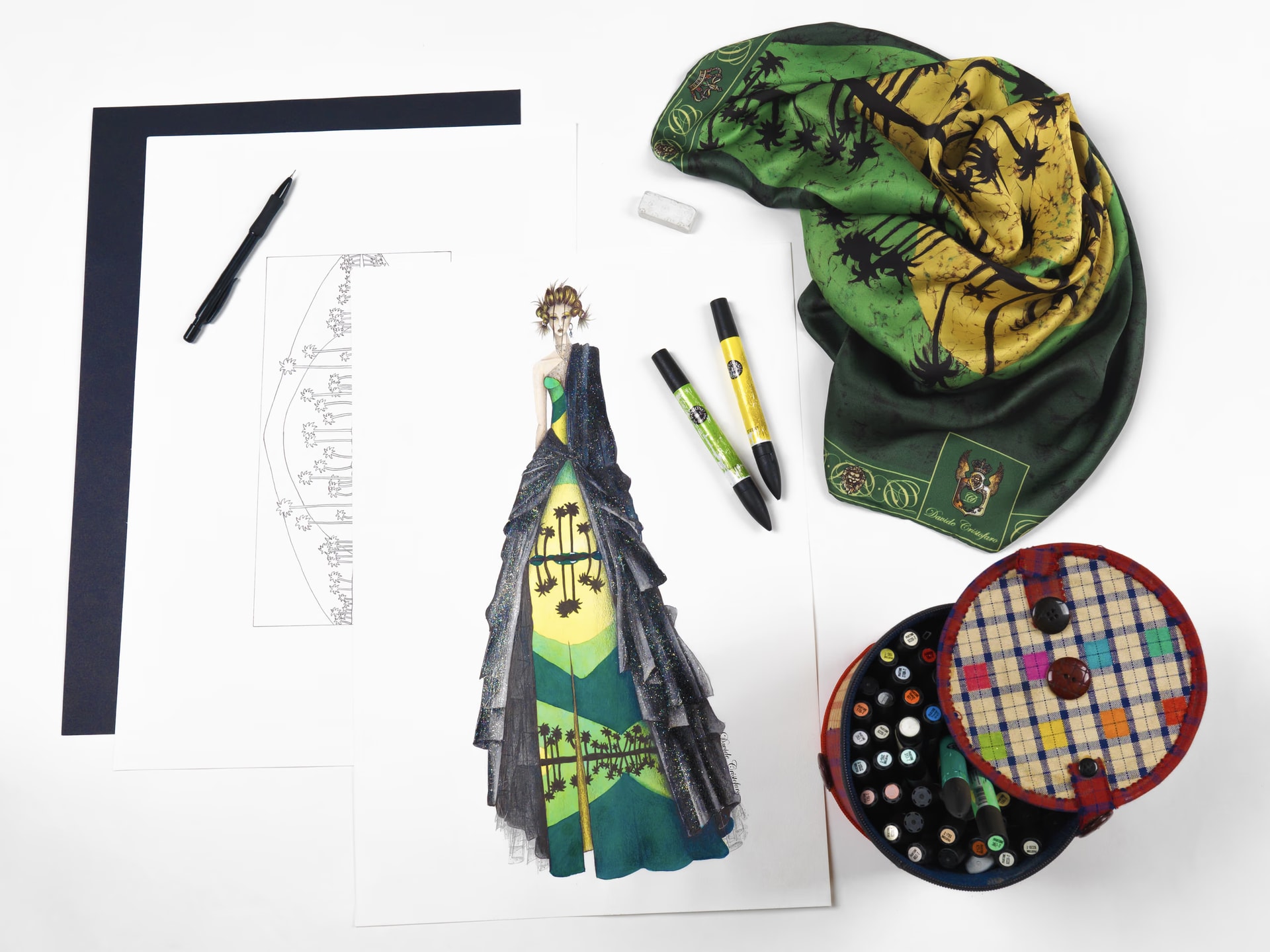Foulard Palm Beach verde e giallo con palme accanto al bozzetto di un abito, pennarelli, una squadra nera e una scatola di bottoni