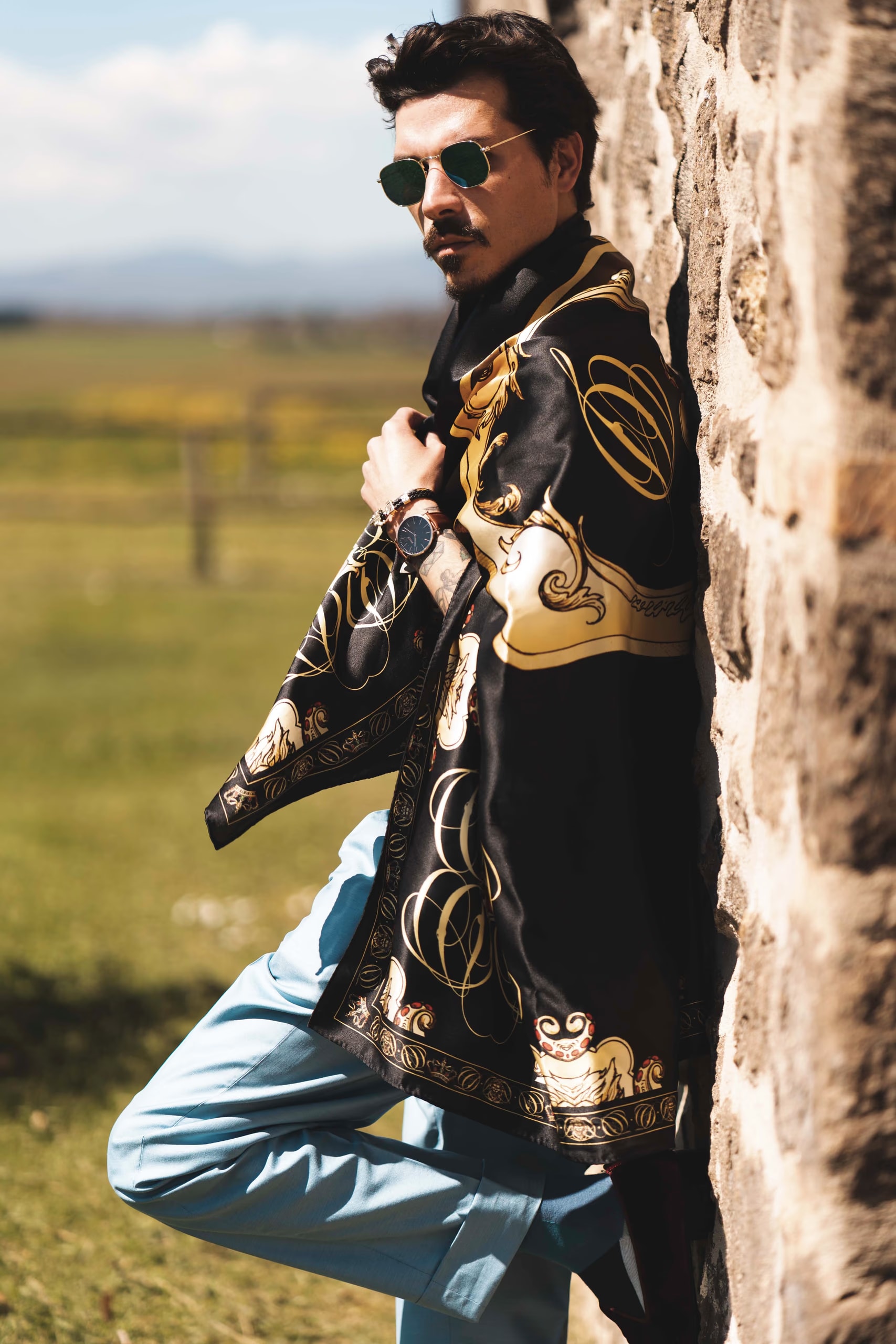 Guglielmo Giovannoni appoggiato a un muro di pietra in campagna, con un foulard di seta nero e oro Davide Cristofaro