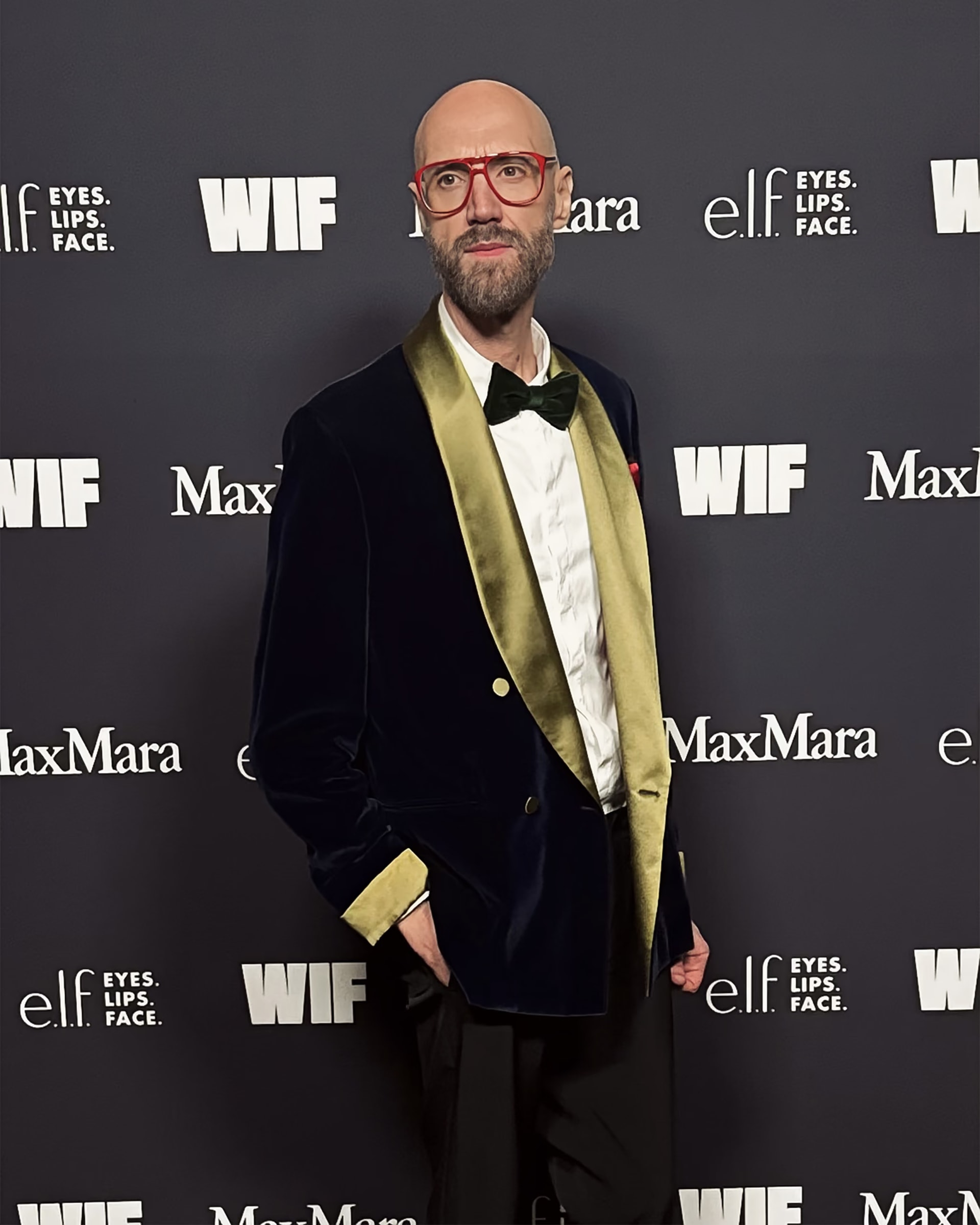 Raimondo Rossi con occhiali rossi, smoking di velluto blu con revers dorati e papillon nero davanti al pannello WIF e Max Mara