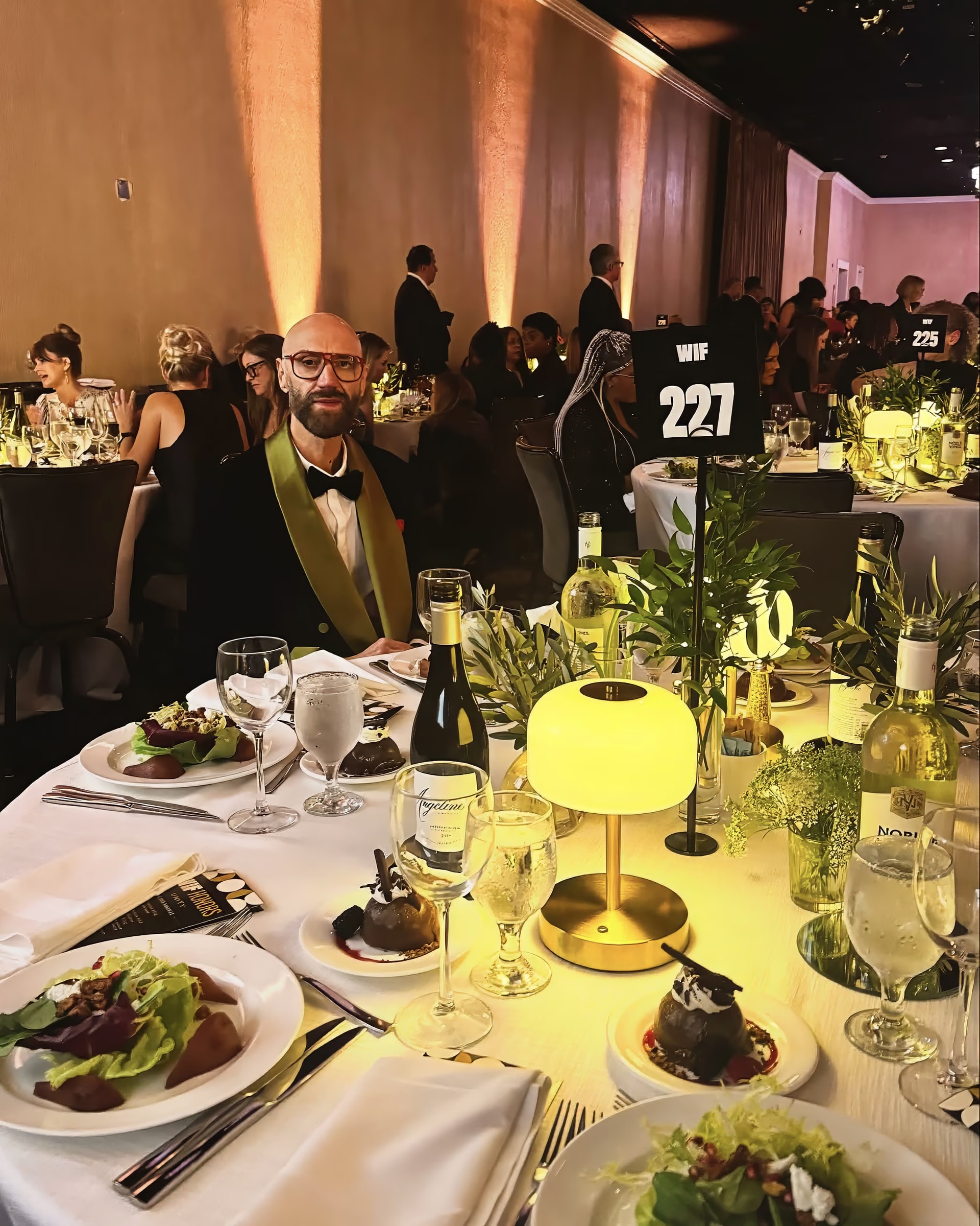 Raimondo Rossi seduto al tavolo 227 del gala WIF Honors 2025, fra lampade dorate, calici e ospiti in sala